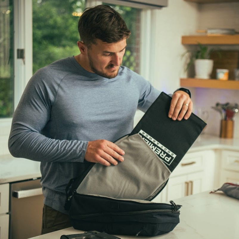 A man in a blue long-sleeve shirt stands in a kitchen, placing the Vertx/Premier Armored Vertx Any Day Backpack Bundle on the counter; a handgun and magazine are visible nearby.