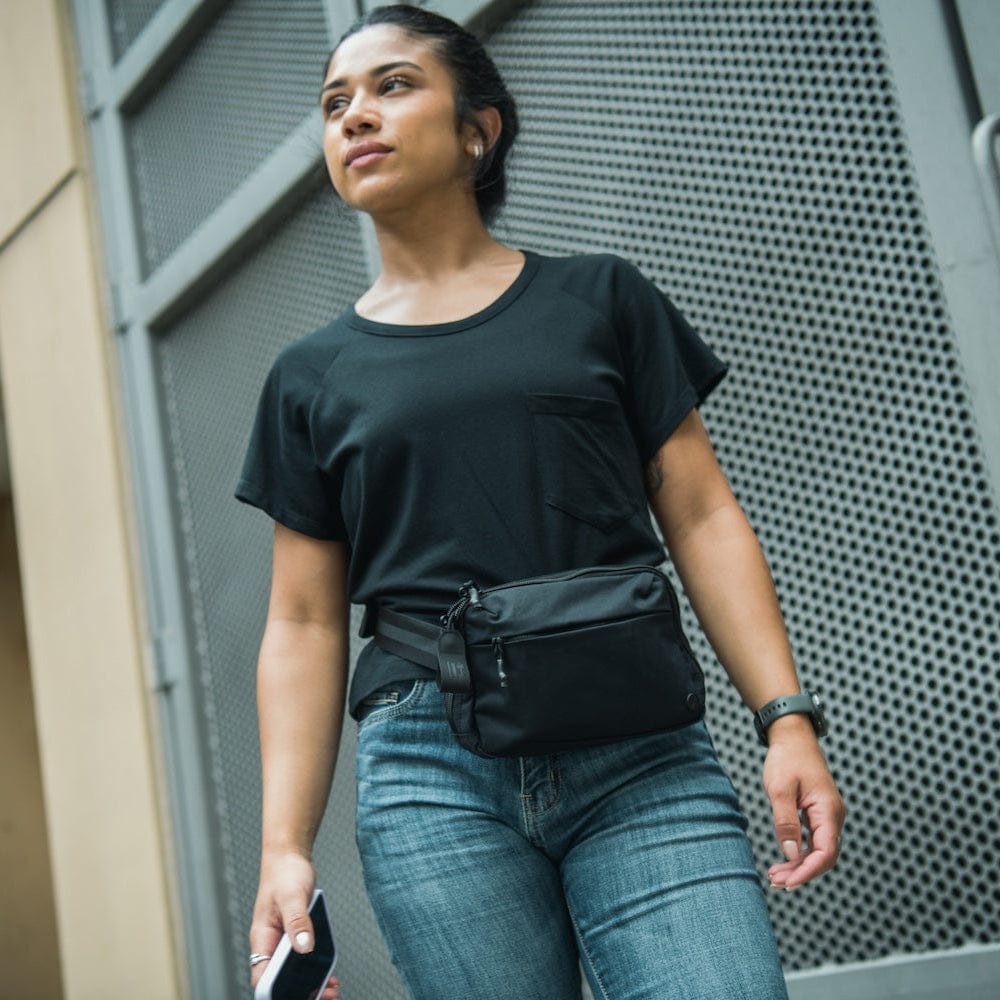 A woman wearing a black t-shirt, jeans, and the Vertx/Premier Armored Vertx Everyday Fanny Pack 2.1 Bundle walks outside by a metal fence, phone in hand, showcasing practical everyday carry style. Another person in a light shirt and cap stands in the background.