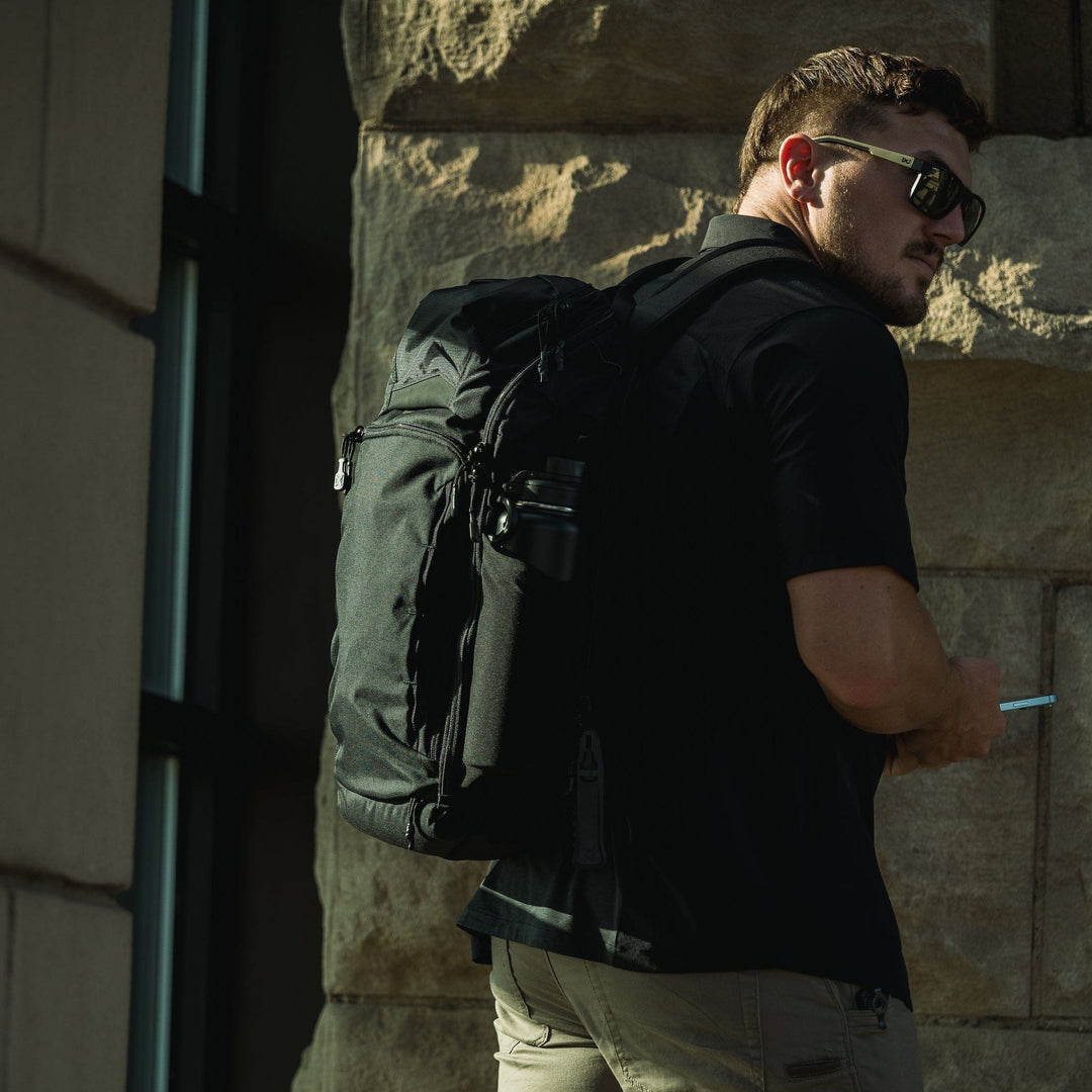A man in sunglasses and a black shirt carries the Vertx Gamut 26L Pack by Vertx, featuring rapid access, while holding a smartphone in front of a stone building, partially turned toward the camera.