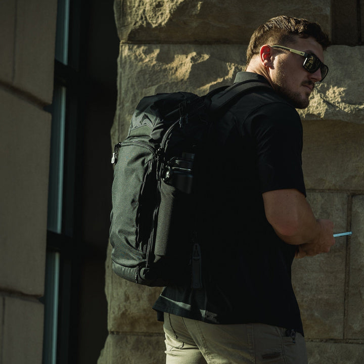 A man in sunglasses and a black shirt carries the Vertx Gamut 26L Pack by Vertx, featuring rapid access, while holding a smartphone in front of a stone building, partially turned toward the camera.