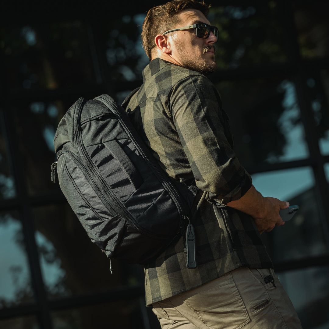 A man in sunglasses, a checkered shirt, and beige pants carries a Vertx Gamut 22L Sling from Vertx over his shoulder and holds a phone while walking past a windowed building in sunlight.