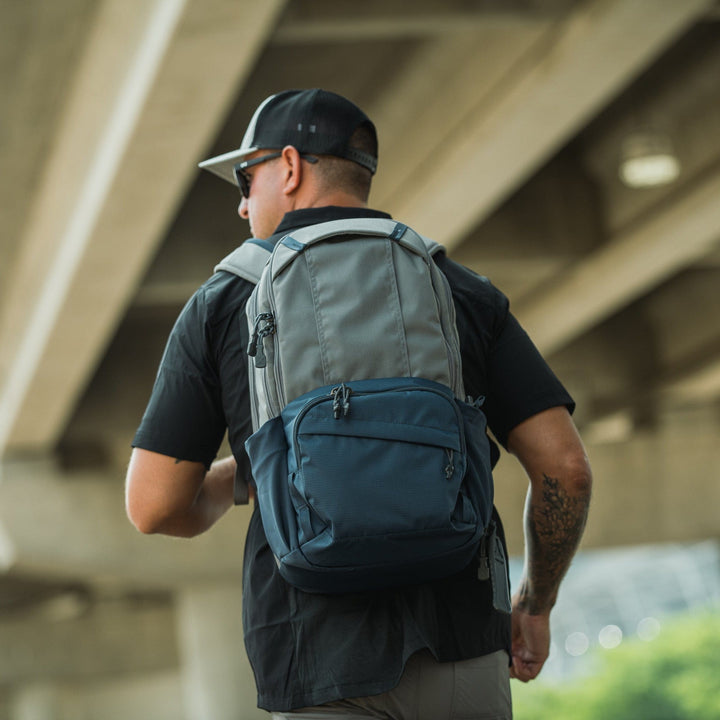 A man wearing a black cap, black shirt, and Vertx Ready Pack X backpack—ideal for everyday carry with its CCW compartment—walks outdoors under a large concrete overpass, with greenery and structure in the background.