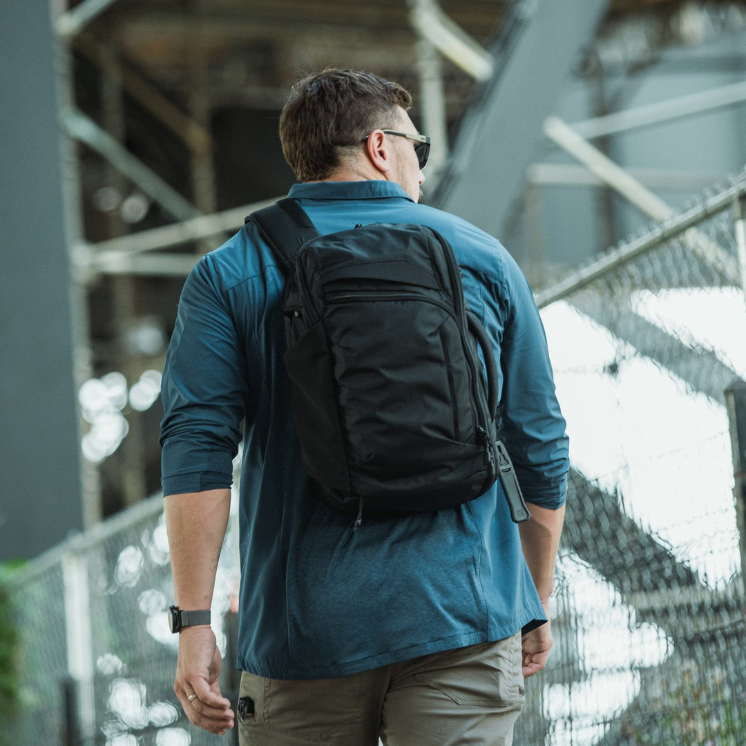 A person in a blue long-sleeve shirt and beige pants walks outdoors, carrying the Vertx Gamut 18L Sling by Vertx over one shoulder. A chain-link fence and industrial structures appear in the background.