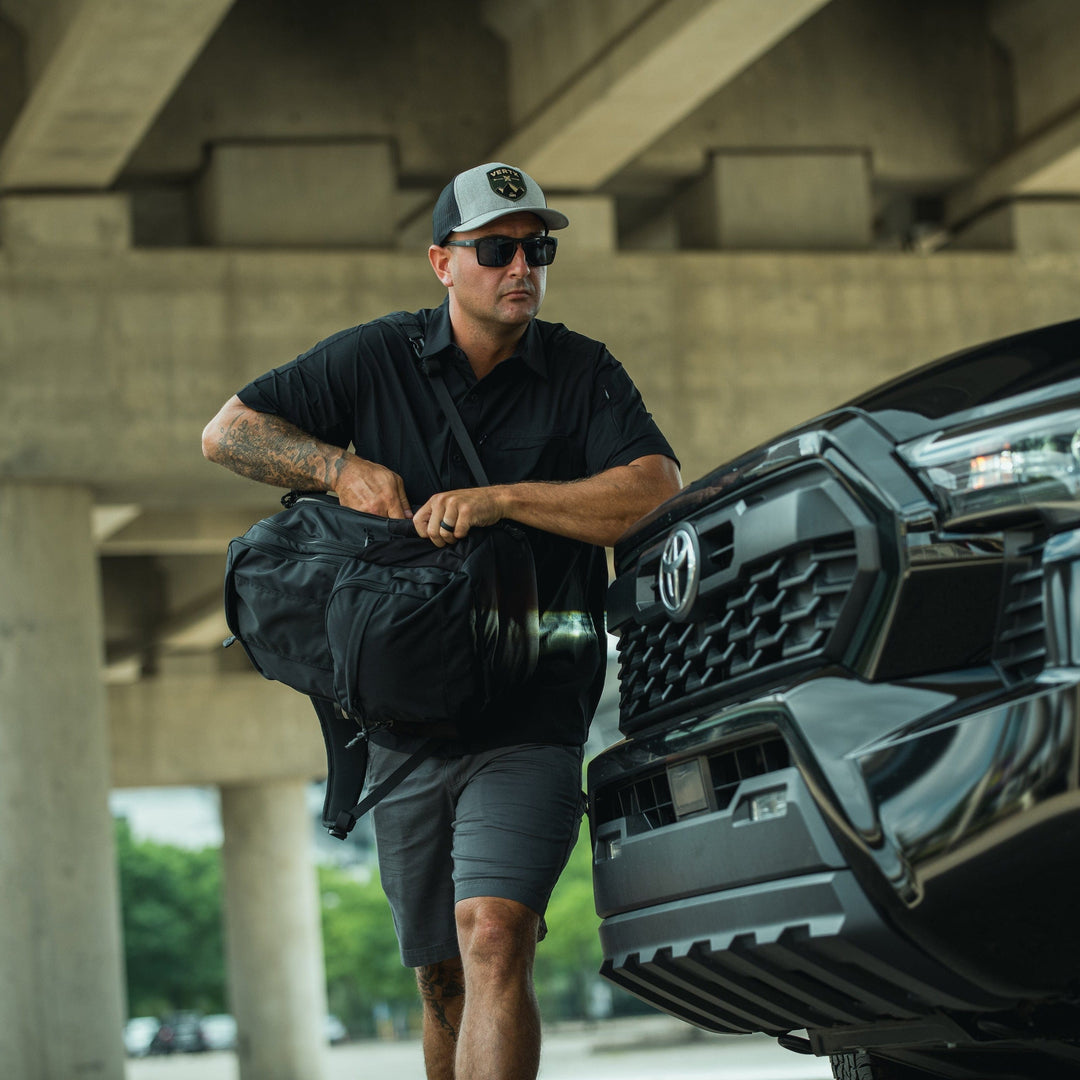 A man in a black shirt, shorts, sunglasses, and a cap pulls a Vertx Ready Pack X with laptop sleeve from a black Toyota truck under a concrete overpass.