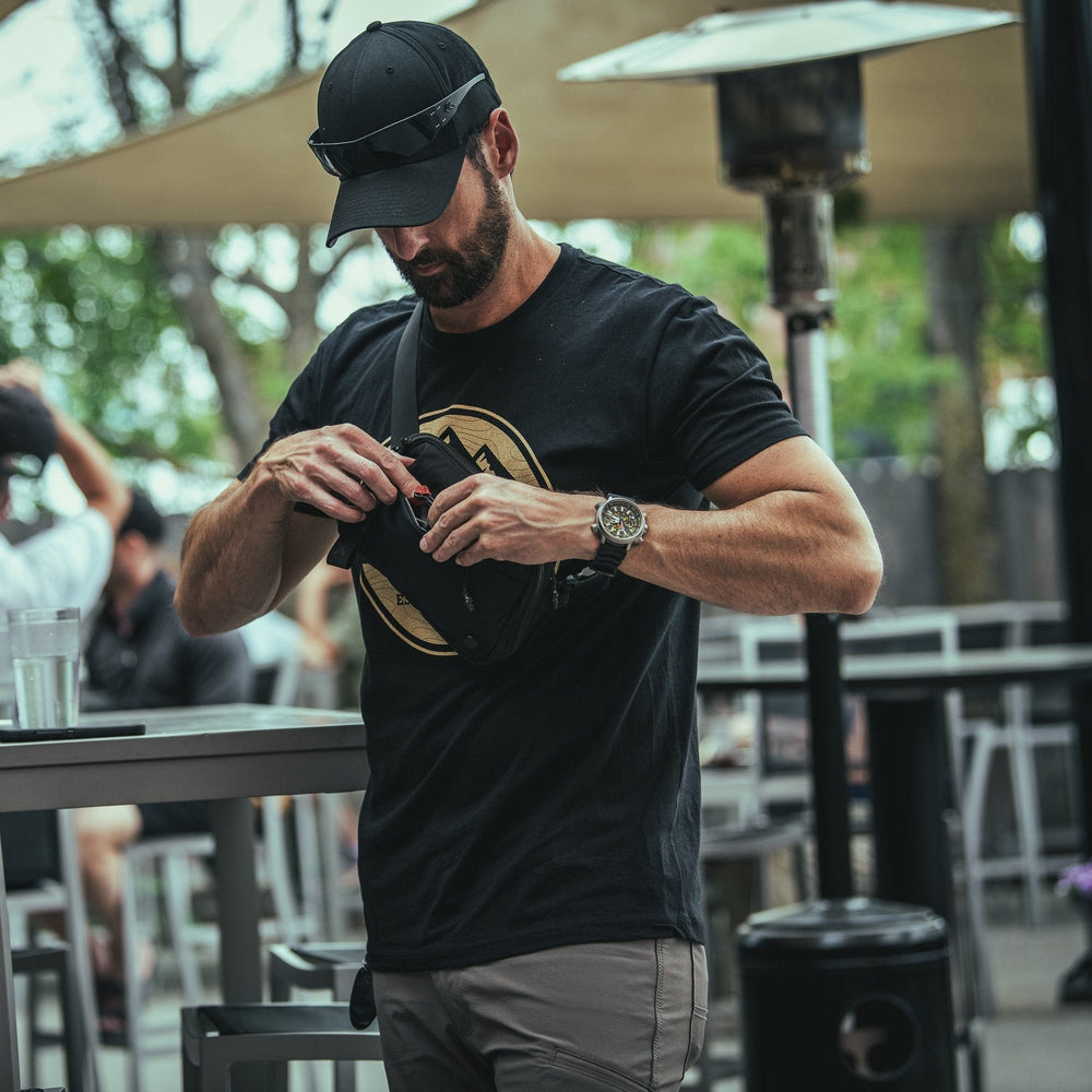 A man in a black cap, sunglasses, and black t-shirt stands outdoors by a table, holding and looking into his Vertx Everyday Fanny Pack 2.1. Other people are seated in the blurred background.