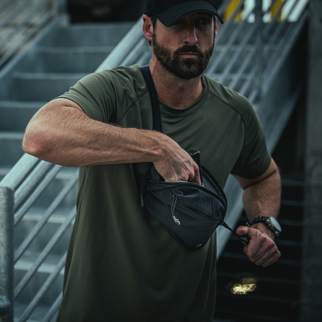 A person in a dark cap and olive-green t-shirt stands on an outdoor staircase, reaching into their Vertx Everyday Fanny Pack. The background consists of metal railings and concrete steps.