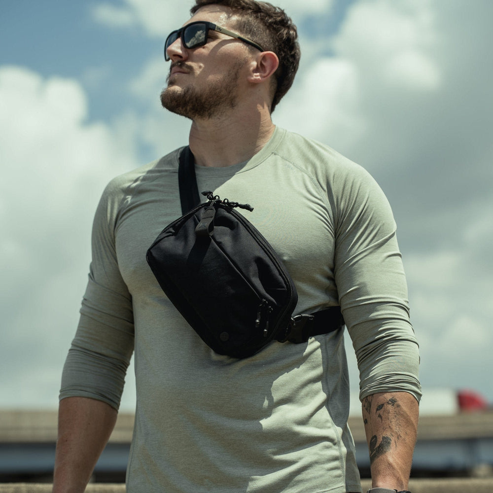 A man stands outdoors under a cloudy sky, wearing sunglasses, a light gray long-sleeve shirt, and the Vertx Everyday Fanny Pack+ by Vertx crossbody. Buildings are faintly visible in the background.