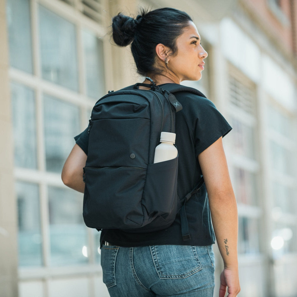 A person with dark hair in a bun, wearing a black t-shirt, jeans, and carrying the Vertx Any Day Backpack by Vertx—with a water bottle in its side pocket—walks down a city sidewalk lined with buildings.
