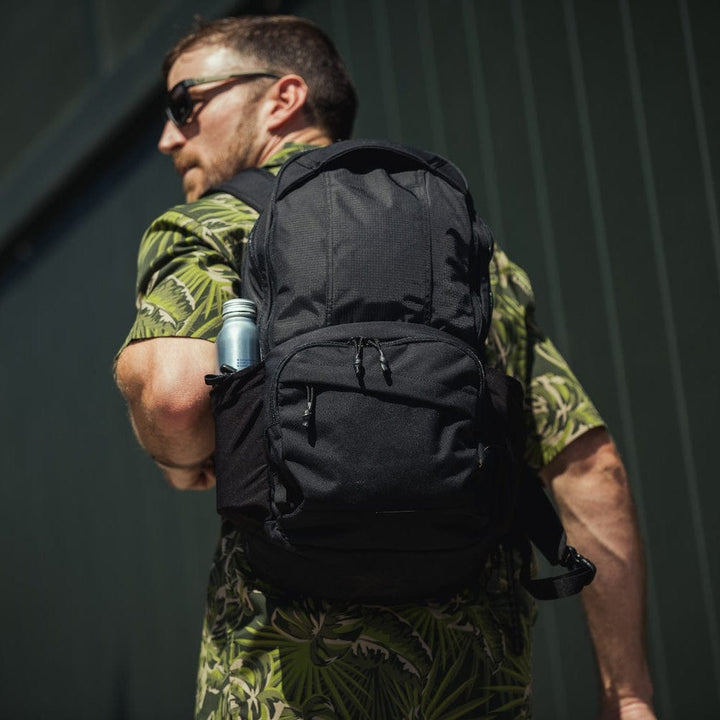 A man in sunglasses and a green patterned shirt carries the Vertx/Premier Armored Vertx Ready Pack X Bundle with a silver can in the side pocket, standing in front of a dark vertical-panel wall.