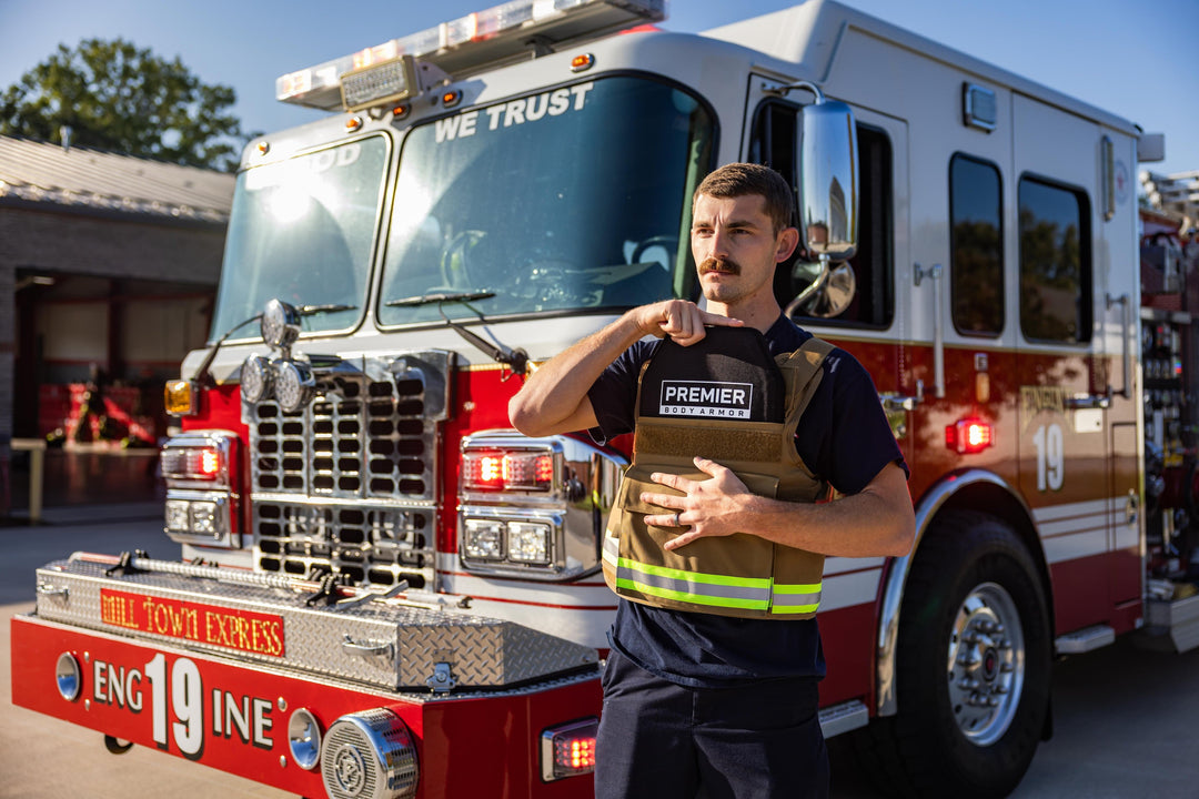 A man standing in front of a red fire truck puts on a protective vest labeled "Premier." The fire truck has the number 19 on it and is parked outside a fire station.
