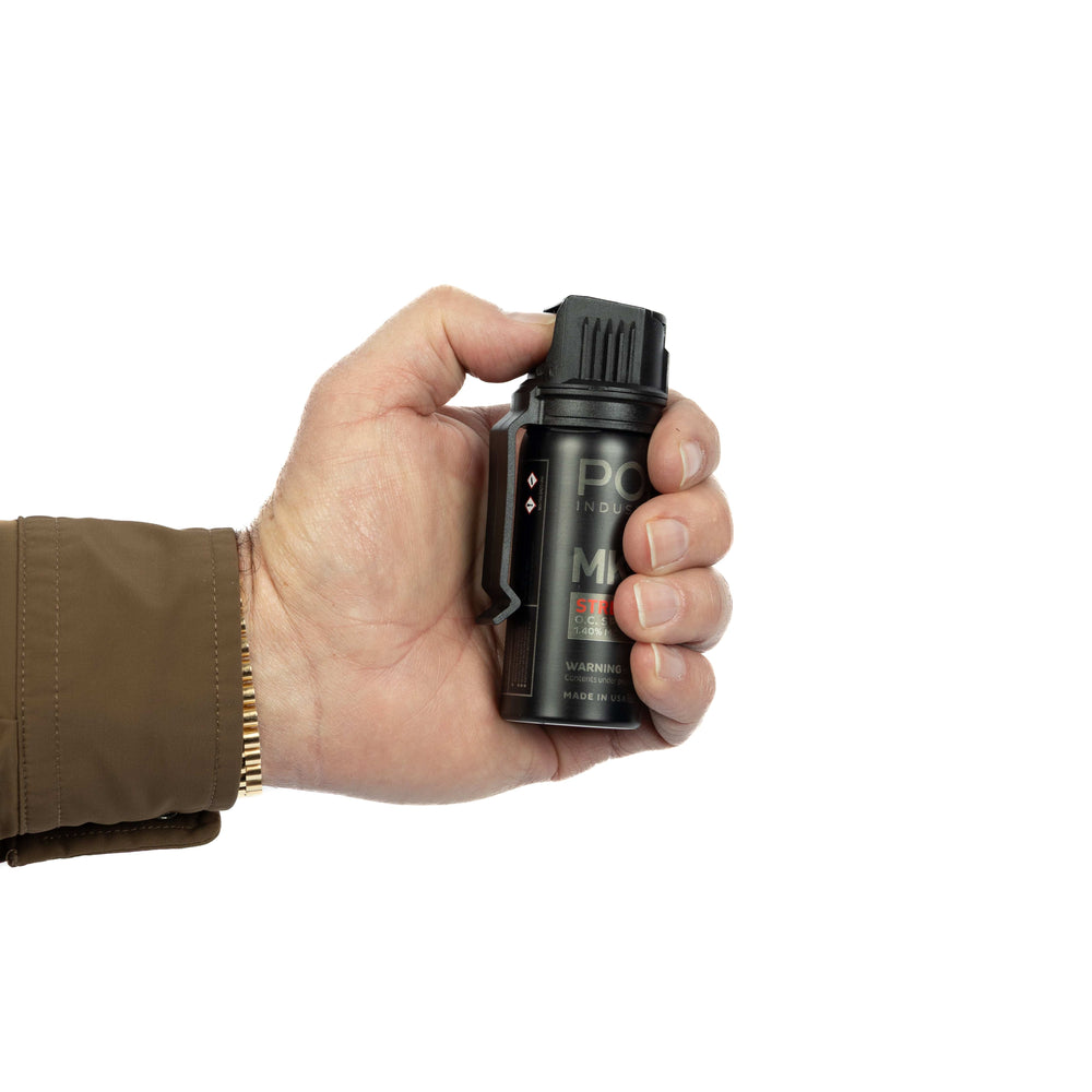 A hand in a brown sleeve and gold bracelet holds the POM MK-3 Pepper Spray by POM, a compact, professional-grade canister with a clip for easy access, shown against a white background.