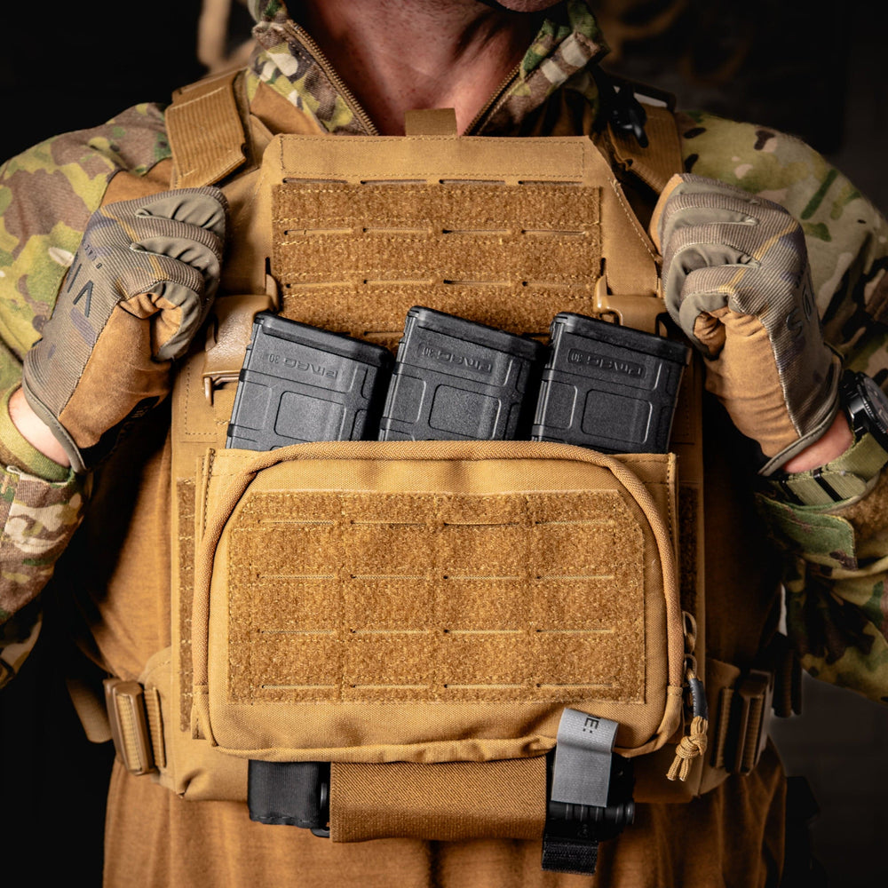 A person wearing camouflage and gloves adjusts a Premier Body Armor Fortis Pro Placard (556/39) with three front AR-15 mag holders featuring secure Kydex retention; the blurred background is dark and out of focus.