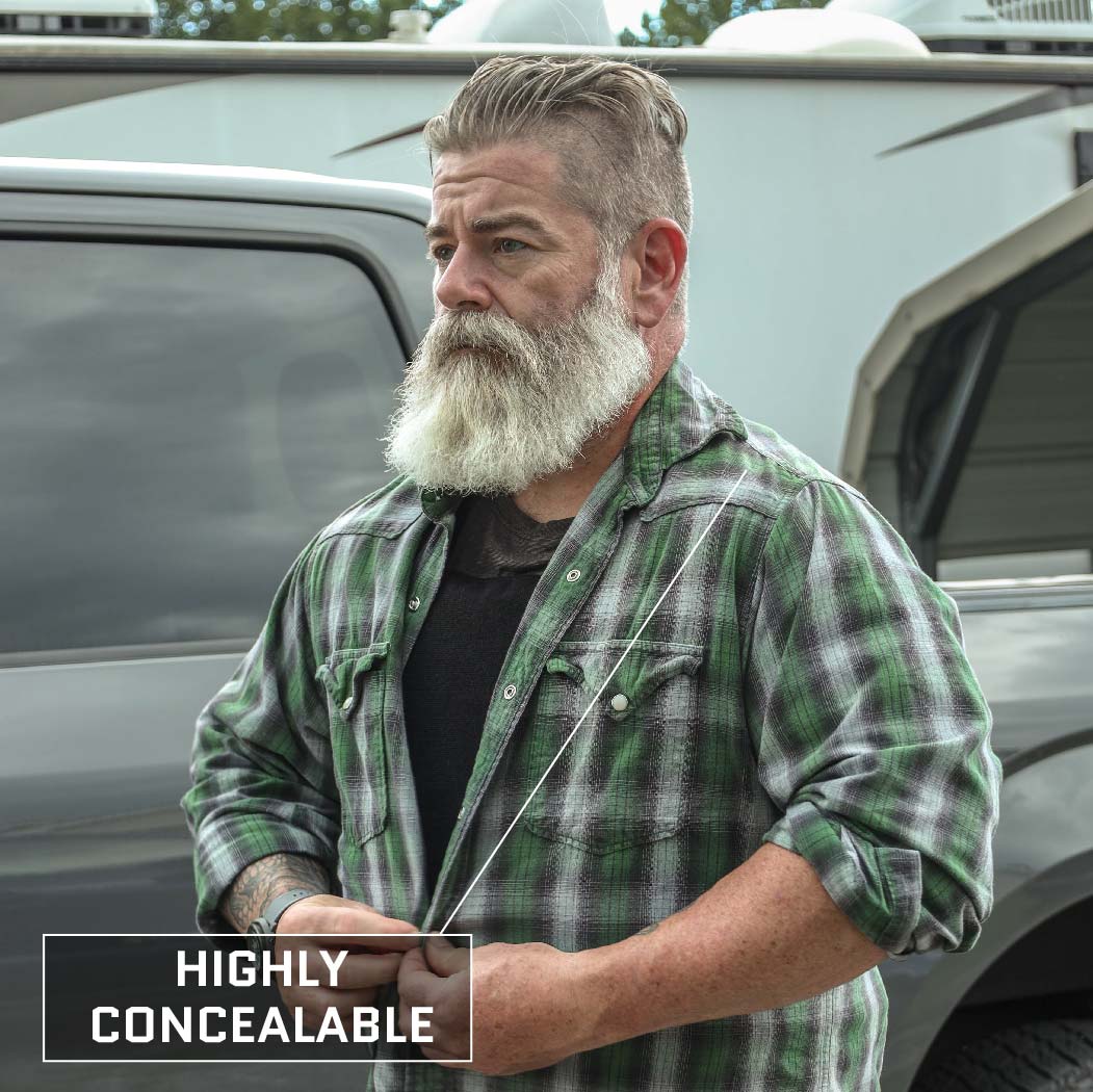 A man with a gray beard and mustache wears a green plaid shirt over a black shirt, standing in front of a gray SUV. The text "HIGHLY CONCEALABLE" is superimposed on the bottom-left corner of the image, suggesting he's wearing a Premier Body Armor Concealable Armor Vest - Level IIIA, made in the USA.