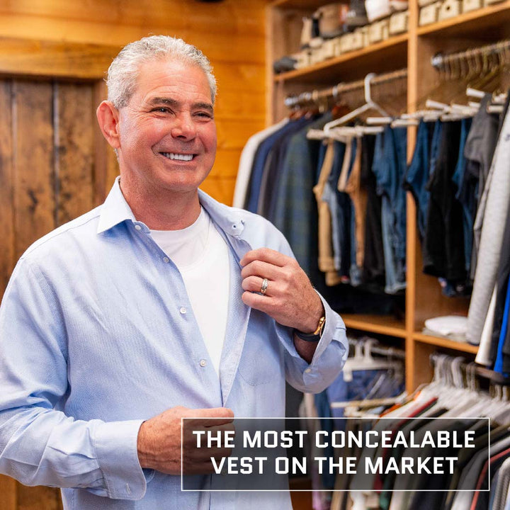 A smiling man in a light blue shirt stands in front of an open closet with clothes hanging on racks. He holds his shirt slightly open, revealing a Premier Body Armor Discreet Executive Vest - Level IIIA underneath. Text on the image reads, "THE MOST CONCEALABLE VEST ON THE MARKET.