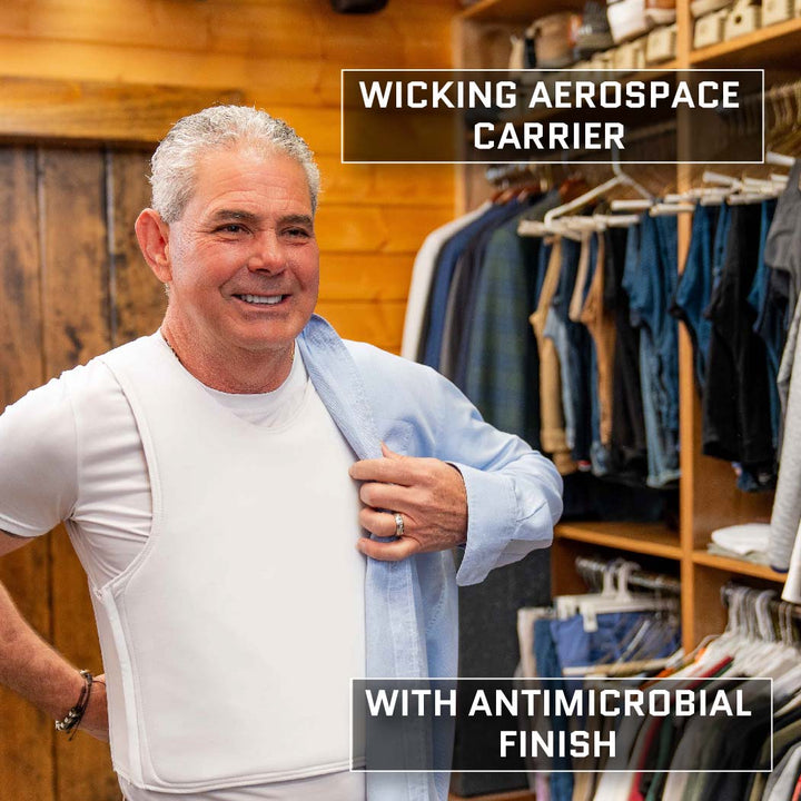 A man is standing in a wooden closet, wearing a white wicking aerospace carrier shirt with an antimicrobial finish and a Premier Body Armor Discreet Executive Vest - Level IIIA. He is smiling and holding a blue shirt slung over his shoulder. Behind him, clothes are hanging on racks.