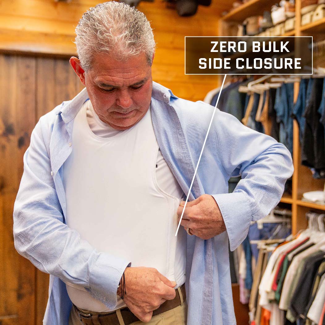 A man is in a wooden-paneled room, putting on a light blue shirt over a white undershirt. Text on the image reads "Zero Bulk Side Closure," with an arrow pointing to the side of the Discreet Executive Vest – Level IIIA from Premier Body Armor. Clothes are hanging on racks in the background.