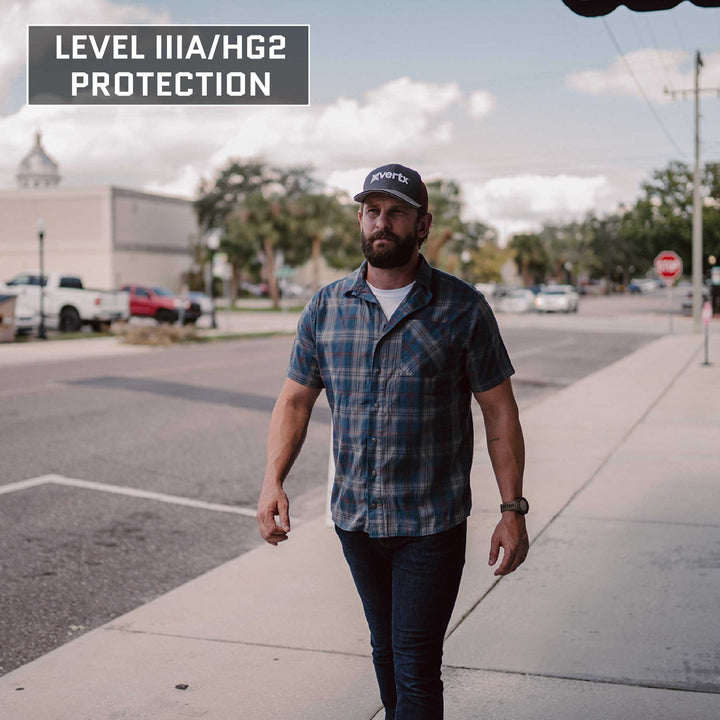 A bearded man wearing a blue plaid shirt, jeans, and a grey cap walks on a sidewalk in an urban area. The text "LEVEL IIIA/HG2 PROTECTION" is positioned at the top left of the image, suggesting he might be wearing Premier Body Armor's Everyday Armor T-Shirt (Armored). Cars and buildings are visible in the background.