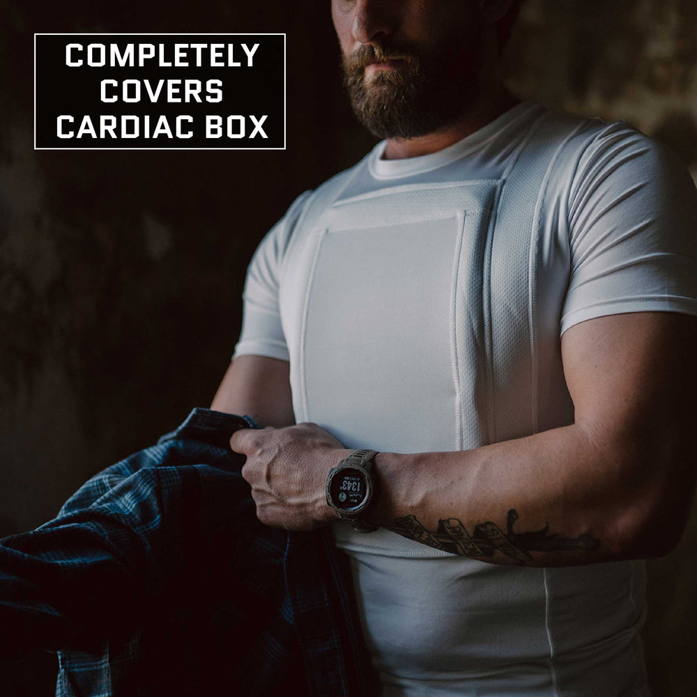 A person wearing a white Premier Body Armor Everyday Armor T-Shirt (Armored) that fully protects the cardiac box area is shown from the chest up. The individual is adjusting or putting on a blue plaid shirt over it. Text in the upper left corner reads, "Completely Covers Cardiac Box.