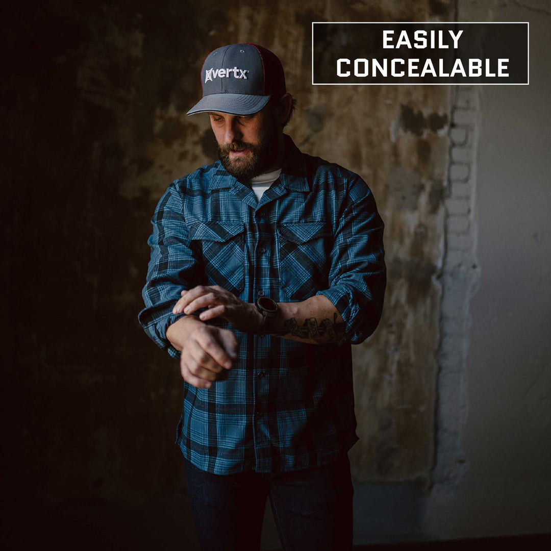 A man wearing a plaid flannel shirt and a cap is adjusting his sleeves while standing against a dark, textured background. The text "easily concealable" is displayed in the top right corner, hinting at the Premier Body Armor Everyday Armor T-Shirt (Armored) he might be discreetly wearing.