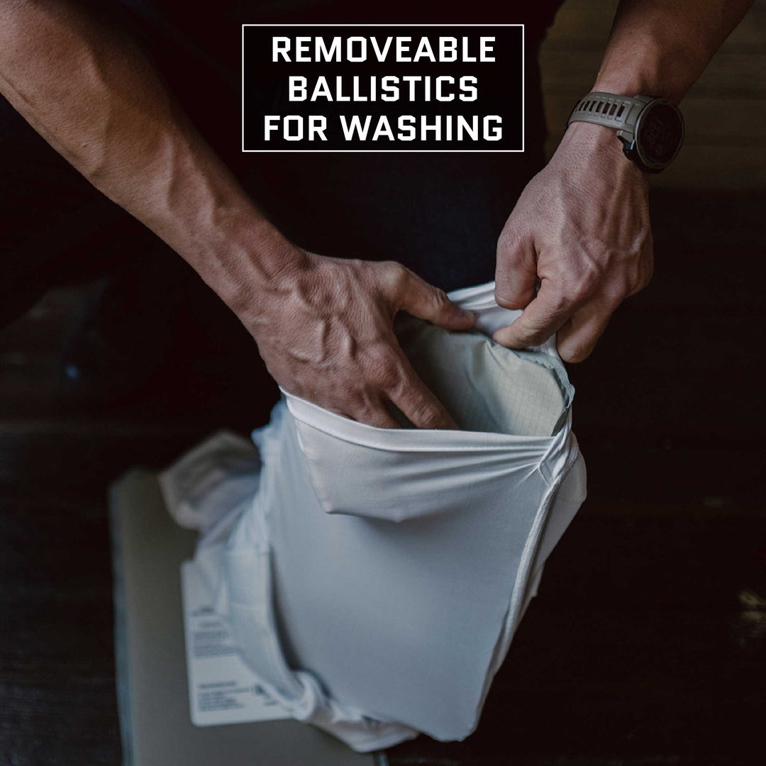 A person is removing ballistic inserts from an Everyday Armor T-Shirt (Armored) by Premier Body Armor. The text on the image reads "REMOVABLE BALLISTICS FOR WASHING." Wearing a wristwatch and standing on a dark-colored surface, the individual demonstrates how easy it is to clean this concealable T-shirt with body armor.