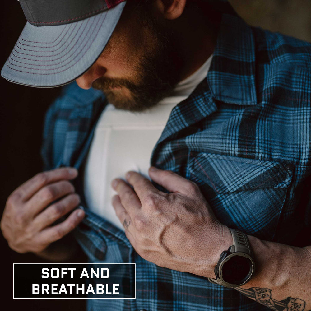 A man wearing a blue plaid shirt and a grey and red cap is pulling his shirt open to reveal the Premier Body Armor Everyday Armor T-Shirt (Armored), a soft and breathable white undershirt that potentially conceals body armor. He has tattoos on his left hand and wrist, complemented by a dark wristwatch. Text at the bottom reads "Soft and Breathable.”