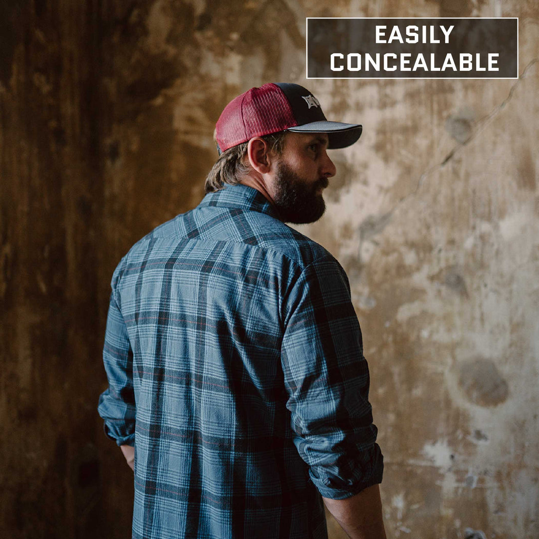 A man with a beard clad in a blue plaid shirt and a red and black cap stands with his back to the camera in a dimly lit room with weathered walls. In the top right corner, the phrase "Easily Concealable" is displayed in a box, subtly referencing Premier Body Armor's Everyday Armor T-Shirt (Armored).