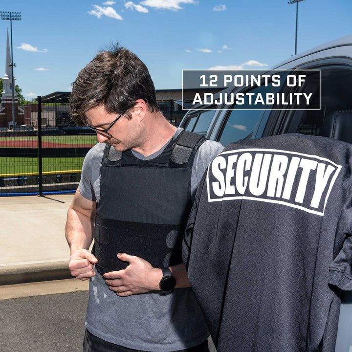 A person in glasses and a gray shirt is adjusting a black Premier Body Armor Overt/Covert Vest Bundle near the side of a vehicle. The vest label on the vehicle reads "12 POINTS OF ADJUSTABILITY." A black garment with "SECURITY" written on it is draped over the vehicle door.