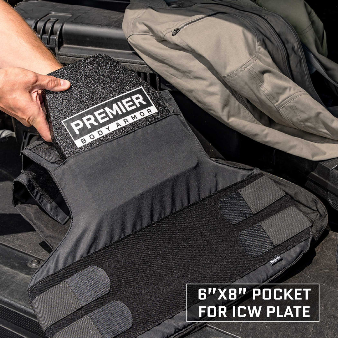 A person is placing a black Premier Body Armor plate into the open pocket of an Overt / Covert Vest Bundle from Premier Body Armor. The pocket, measuring 6 inches by 8 inches, is designed for ICW plates. A backpack sits in the background.