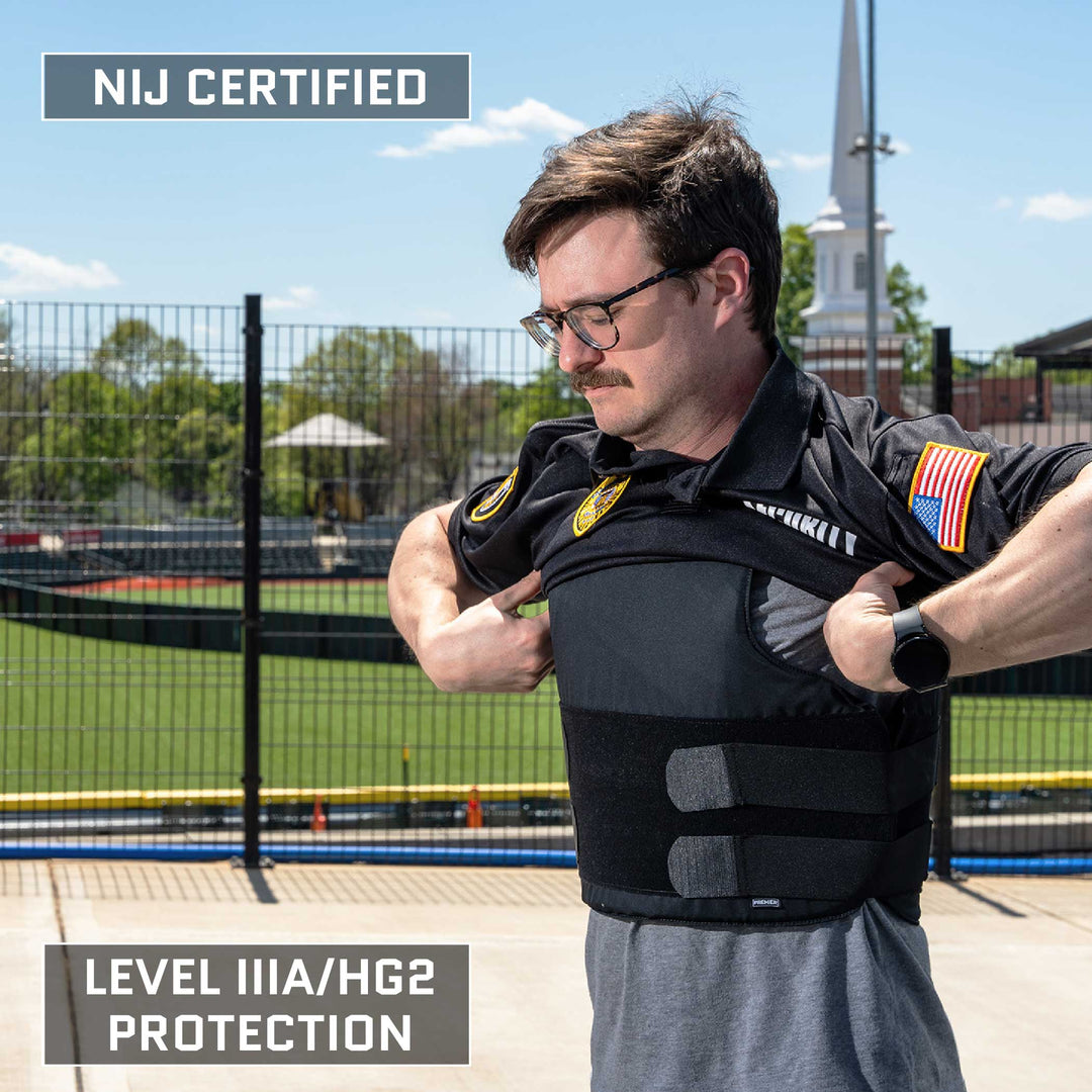 A man with glasses and a mustache is standing outdoors, adjusting a Premier Body Armor Overt / Covert Vest Bundle over his black uniform. The vest has "NIJ Certified" text on it. A field and a building with a white steeple are visible in the background. Text reads "LEVEL IIIA/HG2 PROTECTION.