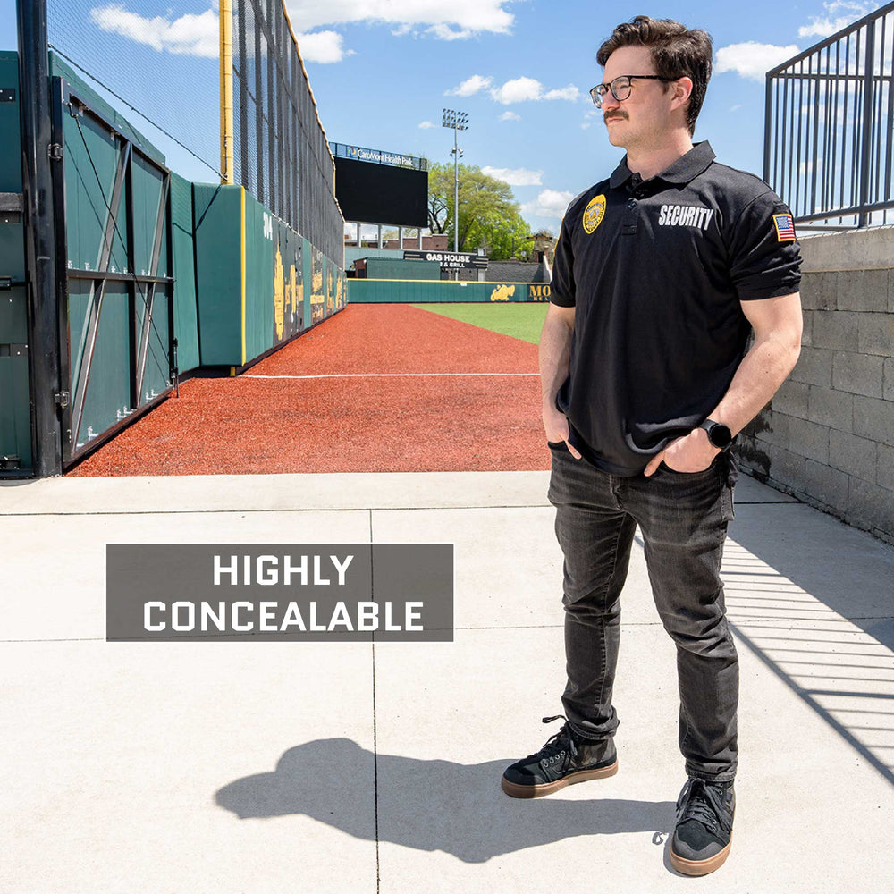 A person stands on a sports field walkway next to a wall, wearing a black "Security" shirt and black pants. The text "Highly Concealable" is displayed near them, promoting the Overt / Covert Vest Bundle from Premier Body Armor. The background includes a green fence, sports field turf, and a blue sky with scattered clouds.