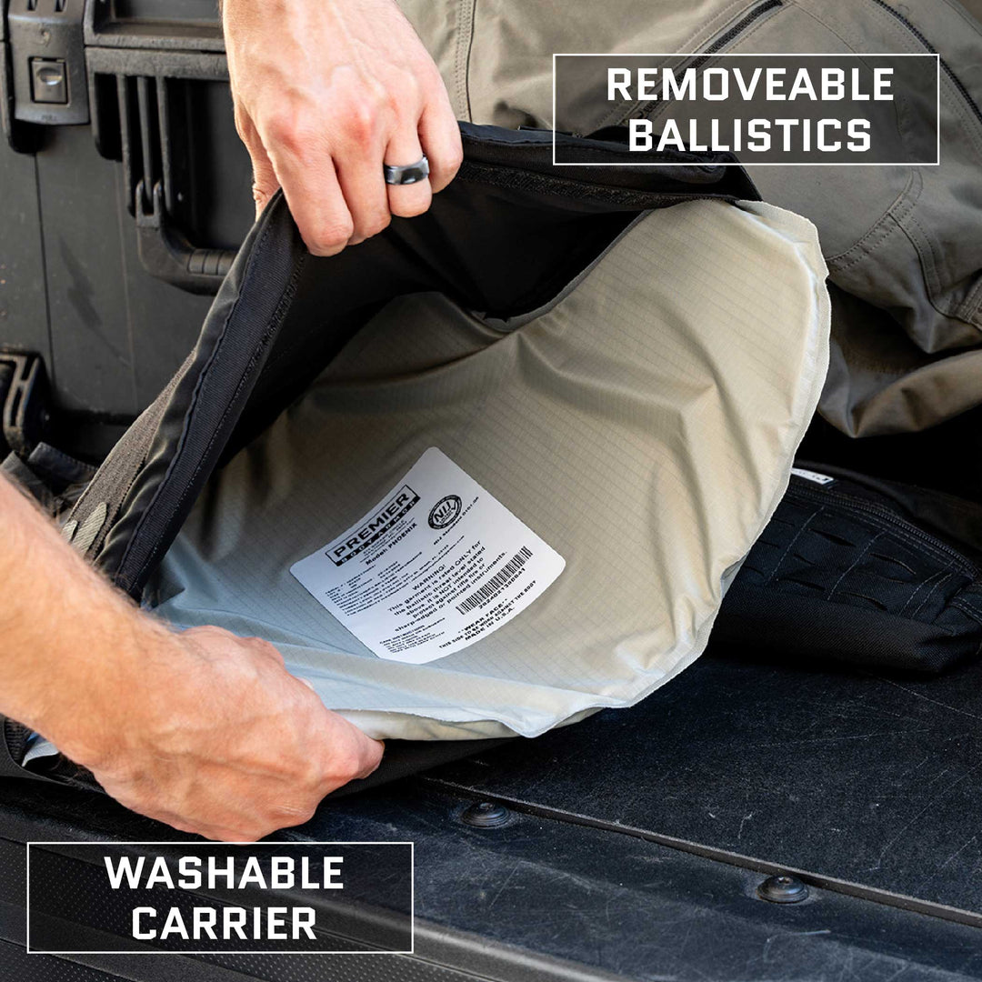 A person is holding open a Premier Body Armor Overt / Covert Vest Bundle (old), revealing the interior label and removable ballistic insert. The vest is marked "Removable Ballistics" and "Washable Carrier." The individual wears a ring, with other tactical equipment in the background.