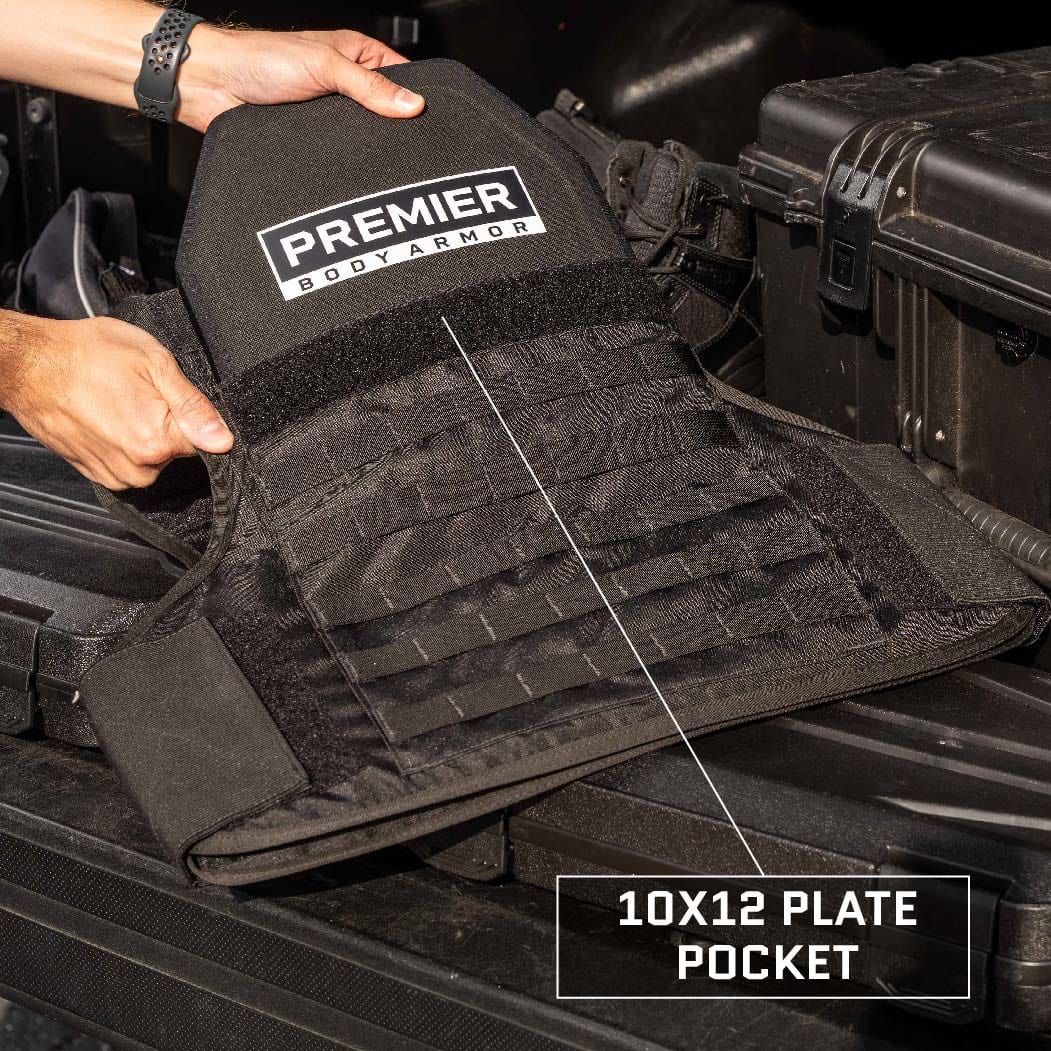 A person holding a black Premier Body Armor Overt/Covert Vest Bundle (old) with a 10x12 plate pocket. The vest is being displayed over a black surface near a protective carrying case.