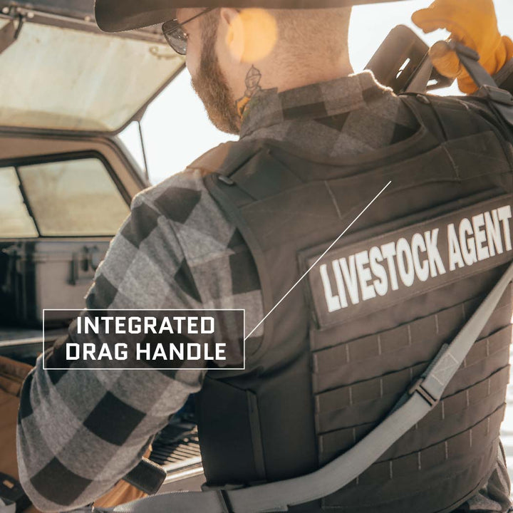 A person in a checkered shirt and black hat is shown from behind, wearing an Overt / Covert Vest Bundle (old) by Premier Body Armor, labeled "LIVESTOCK AGENT." A label indicates an integrated drag handle on the vest. The person stands in front of an open vehicle.