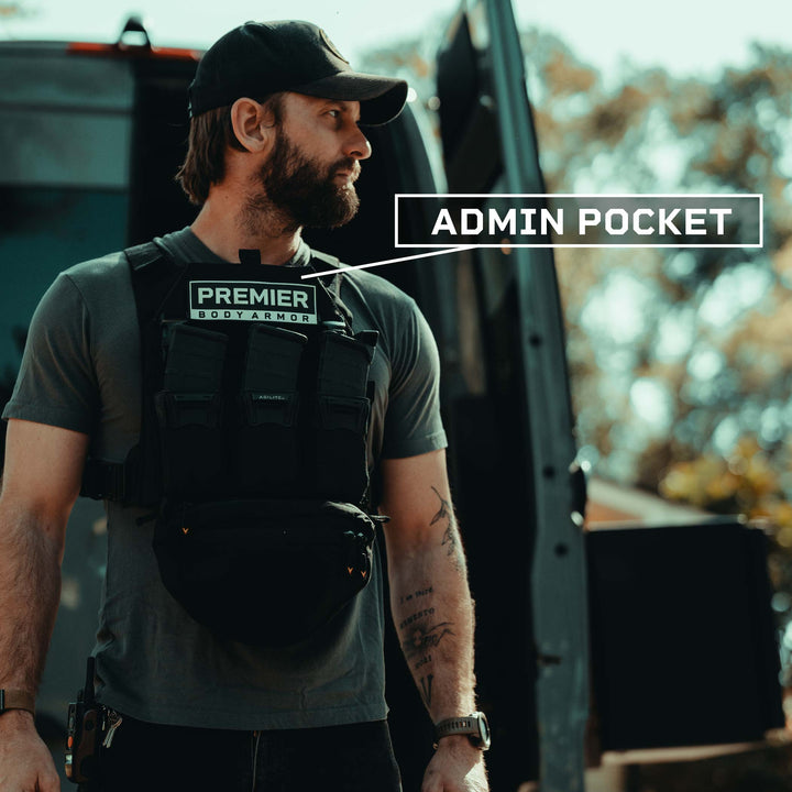 A man wearing a baseball cap and a Fortis Level IV Loadout tactical vest from Premier Body Armor stands next to a vehicle. The vest, equipped with Level IV Plates, features a patch labeled "ADMIN POCKET" with an arrow pointing to a pocket. The background contains blurred greenery and trees.