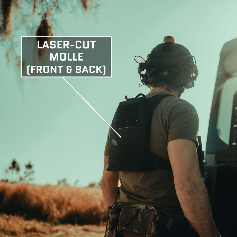 A person wearing a tactical helmet and a Premier Body Armor Fortis Level IV Loadout with laser-cut MOLLE stands near a vehicle, against the backdrop of a natural landscape. The vest, known for its tactical flexibility, features Level IV Plates and laser-cut MOLLE on both the front and back.