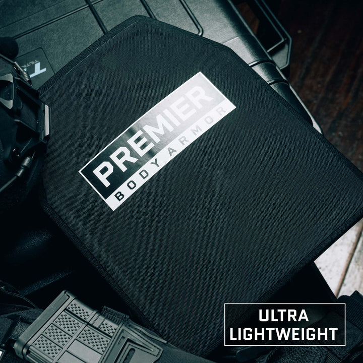 A piece of body armor labeled "Premier Body Armor" rests on a dark surface, featuring the words "Ultra Lightweight" in the lower right corner. Nearby, tactical equipment, including a vest and gun magazine, complements the Fortis III+/RF1 Multi-Curve Level III ballistic plate crafted from ultra-high molecular weight polyethylene.