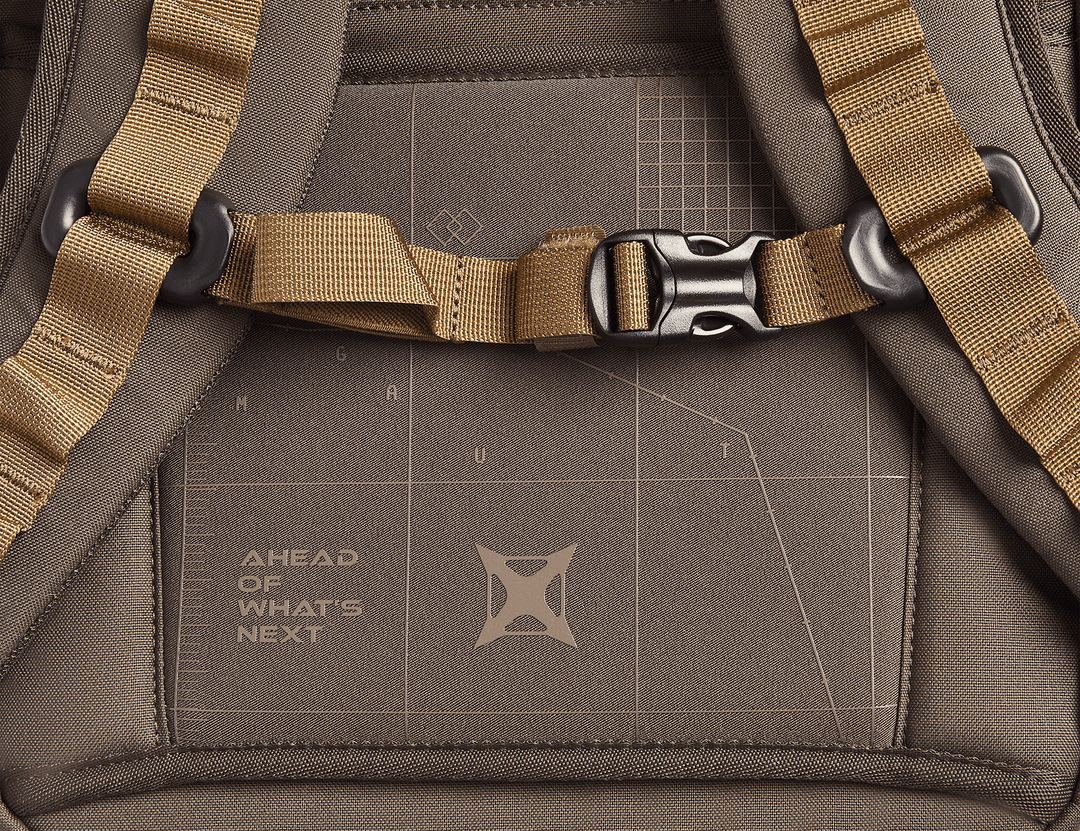 Close-up of the Vertx Gamut 26L Pack, a brown EDC backpack by Vertx, featuring tan straps with a black buckle. The back panel shows a geometric pattern and "AHEAD OF WHAT'S NEXT" printed for quick access to essentials.