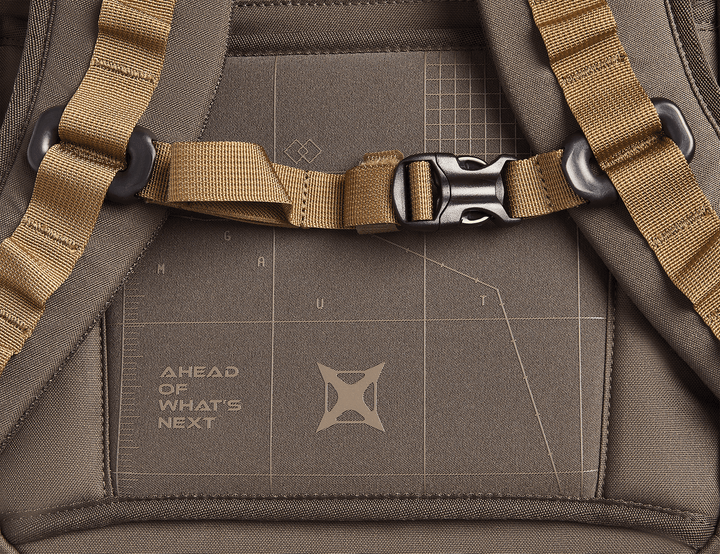 Close-up of the Vertx Gamut 26L Pack, a brown EDC backpack by Vertx, featuring tan straps with a black buckle. The back panel shows a geometric pattern and "AHEAD OF WHAT'S NEXT" printed for quick access to essentials.