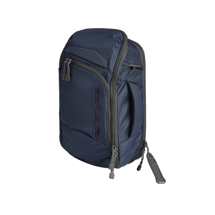The Vertx Gamut 18L Sling by Vertx, a navy blue compact backpack with multiple zippered compartments and gray zipper pulls, stands upright against a plain white background.