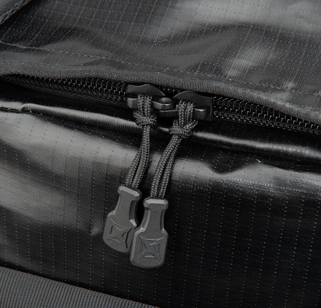 A close-up of a black fabric duffel bag highlights the sturdy design of the Vertx RLT Duffel. The zipper is equipped with two black textured pull tabs, and the material exhibits a subtle grid pattern that allows for customizable storage solutions, enhancing its versatility.