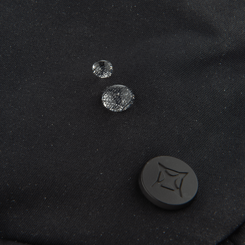 Three water droplets rest on the black fabric of the Vertx Everyday Fanny Pack 2.0, showcasing its water-resistant design by Vertx—making it perfect for off-body concealed carry. A black circular button with a geometric pattern appears at the lower right.