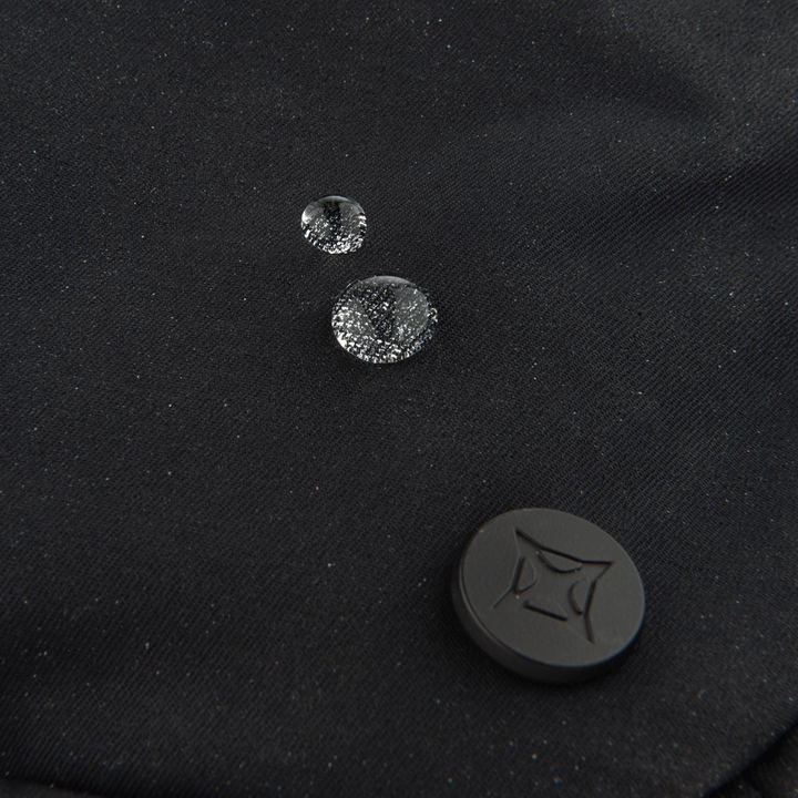 Three water droplets rest on the black fabric of the Vertx Everyday Fanny Pack 2.0, showcasing its water-resistant design by Vertx—making it perfect for off-body concealed carry. A black circular button with a geometric pattern appears at the lower right.