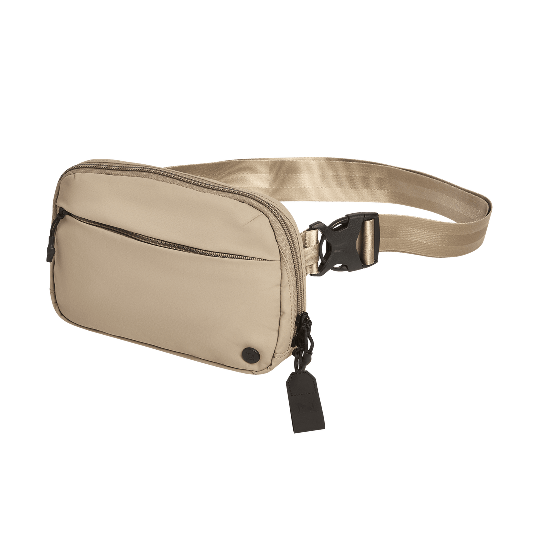 The Vertx Everyday Fanny Pack 2.1 is a beige EDC waist pack with a front zip pocket, adjustable strap with black buckle clasp, small black circular detail, and a black tag on the zipper for added utility.