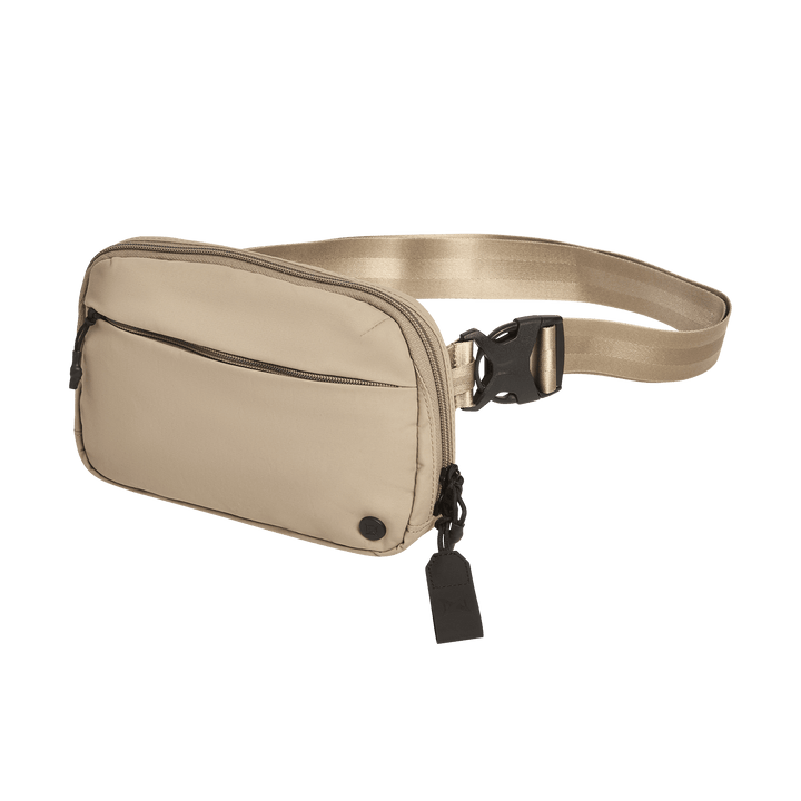 The Vertx Everyday Fanny Pack 2.1 is a beige EDC waist pack with a front zip pocket, adjustable strap with black buckle clasp, small black circular detail, and a black tag on the zipper for added utility.