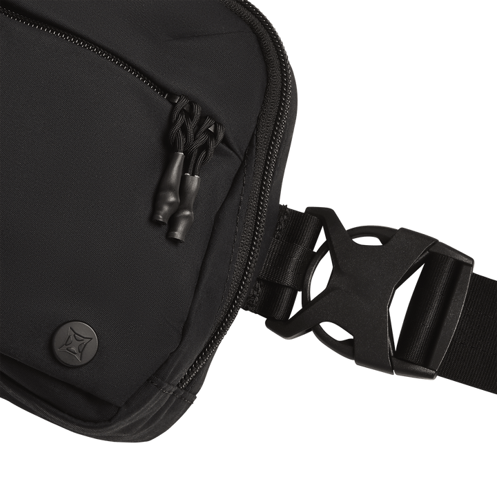 Close-up of the Vertx Everyday Fanny Pack+ by Vertx, featuring a black design with a zippered pocket, double zipper pulls, round Vertx logo, and plastic side-release buckle on a wide strap against a white background.