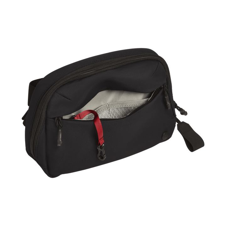 A black Vertx Everyday Fanny Pack+ by Vertx is shown with its main compartment partially open, displaying a gray lining, mesh pocket, and red strap inside. This pouch is ballistic-panel compatible and features a pull tab plus a front zipper.