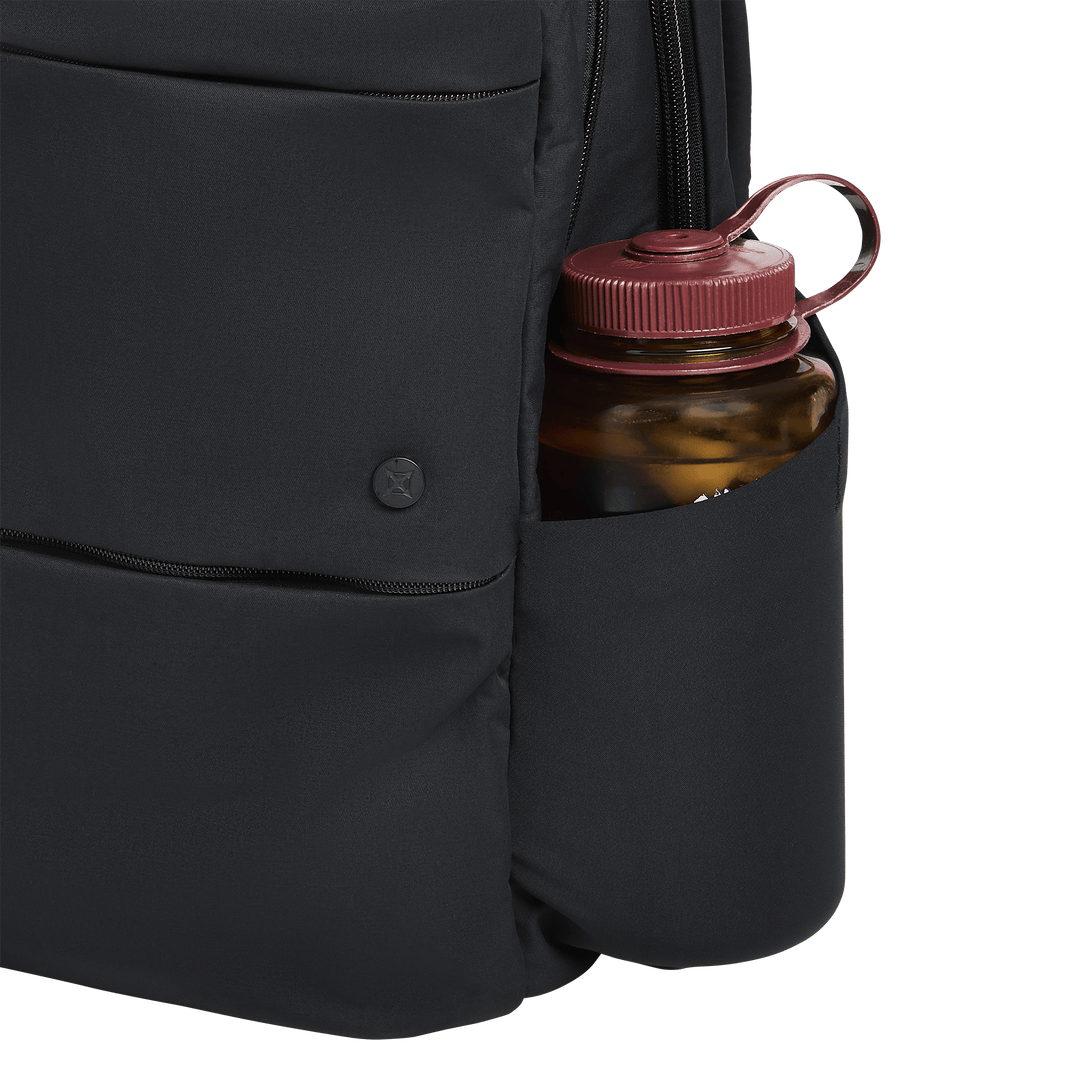 A close-up of the Vertx Any Day Backpack by Vertx, featuring a black design with a red-capped water bottle in its side pocket, set against a white background.