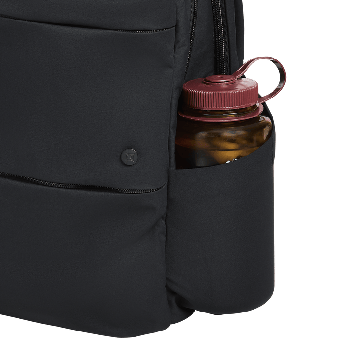 A close-up of the Vertx Any Day Backpack by Vertx, featuring a black design with a red-capped water bottle in its side pocket, set against a white background.