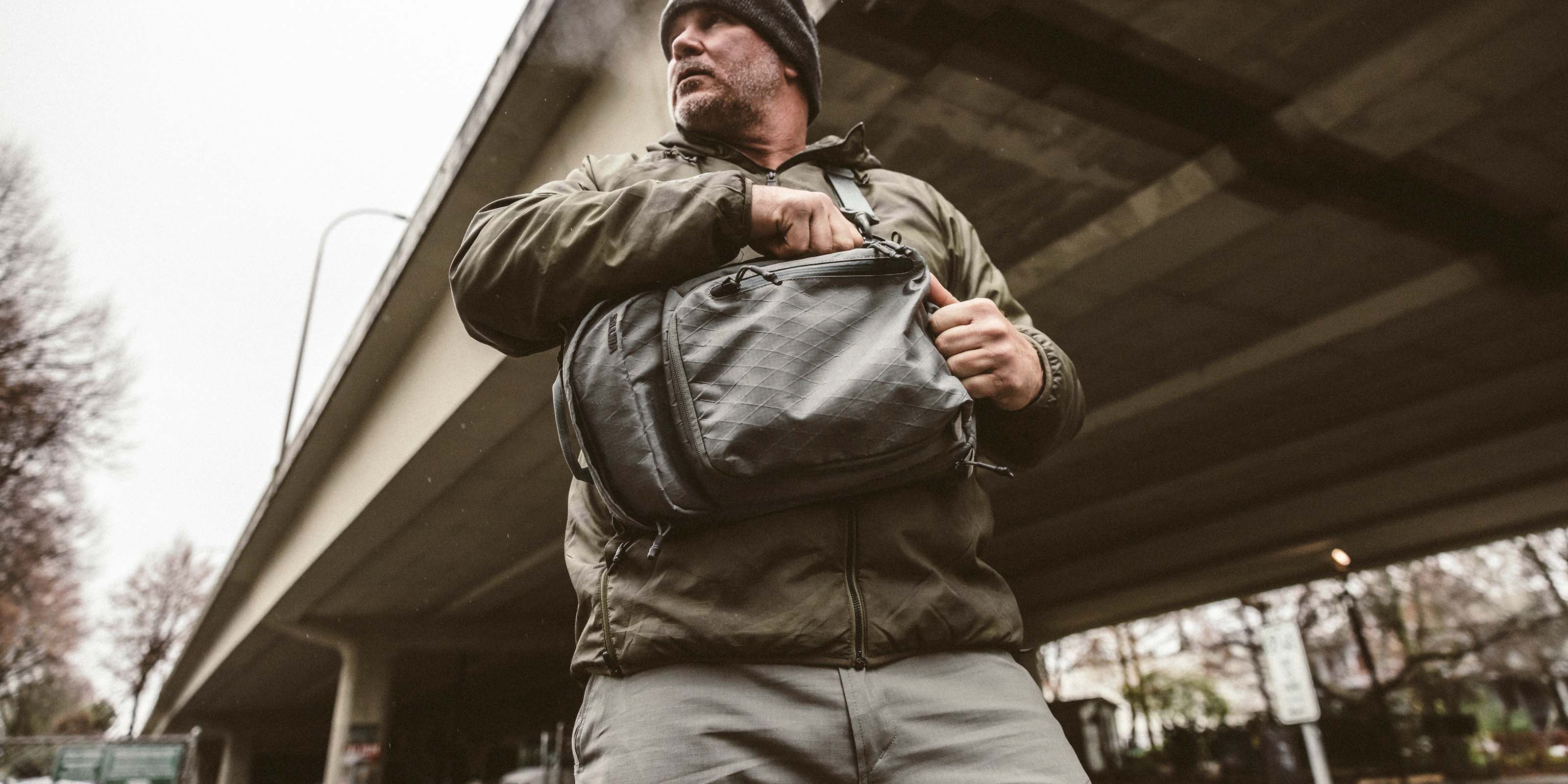 A person stands under an overpass wearing a dark beanie, green jacket, and light-colored pants. They hold a Viktos Upscale Sling XL with one hand on the strap, and their head is turned to the side. The background includes trees and parts of the overpass structure.