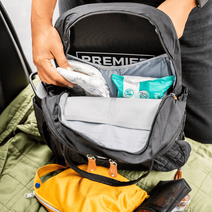 A person is holding open a black backpack, revealing its contents: diapers, a folded cloth, and a grey and white patterned item. In front of the backpack are a yellow bag and a small red bag. A green quilted blanket lies underneath everything. The setup also includes the discrete Bulletproof Diaper Bag Insert Level IIIA from Premier Body Armor for added safety.